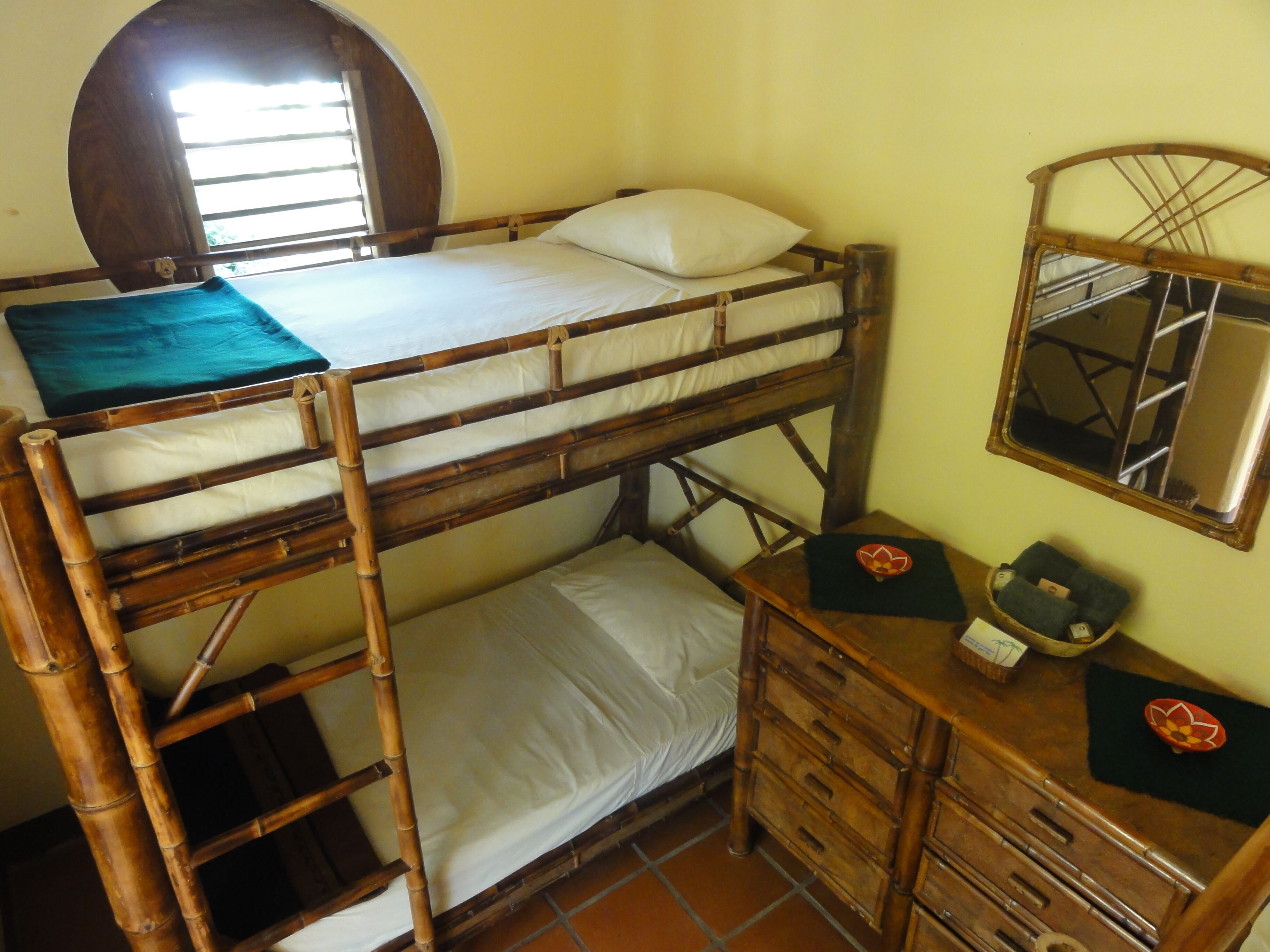 A bunk bed with a wooden frame, a small bedside table with drawers, a mirror, and a window with blinds.