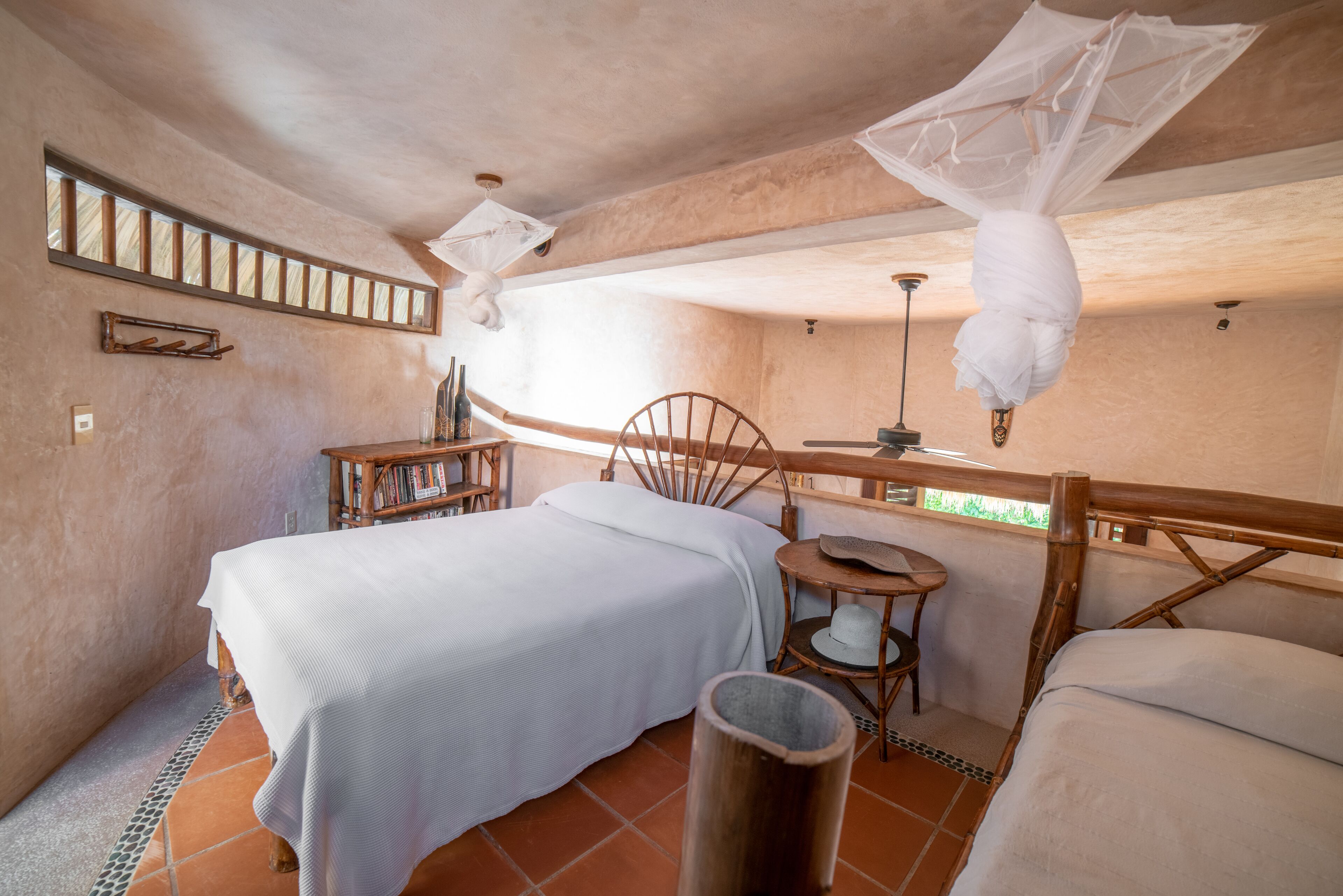 Ein Schlafzimmer mit Bett, kleinem Tisch, Stuhl, einem hängenden Moskitonetz und einem Fenster mit Blick auf Grünflächen.