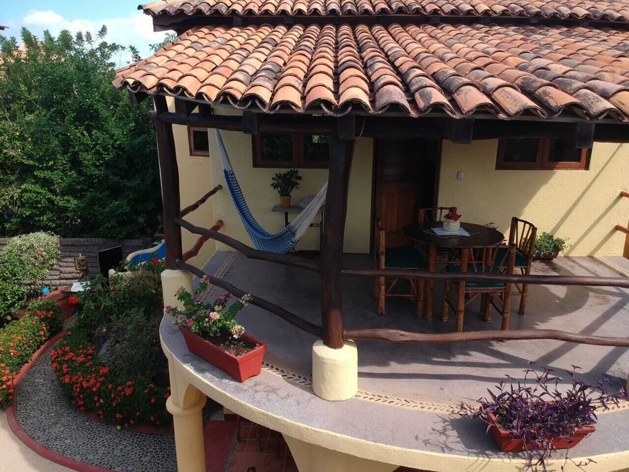 A covered outdoor patio with a dining table and chairs, surrounded by potted plants and a hammock.