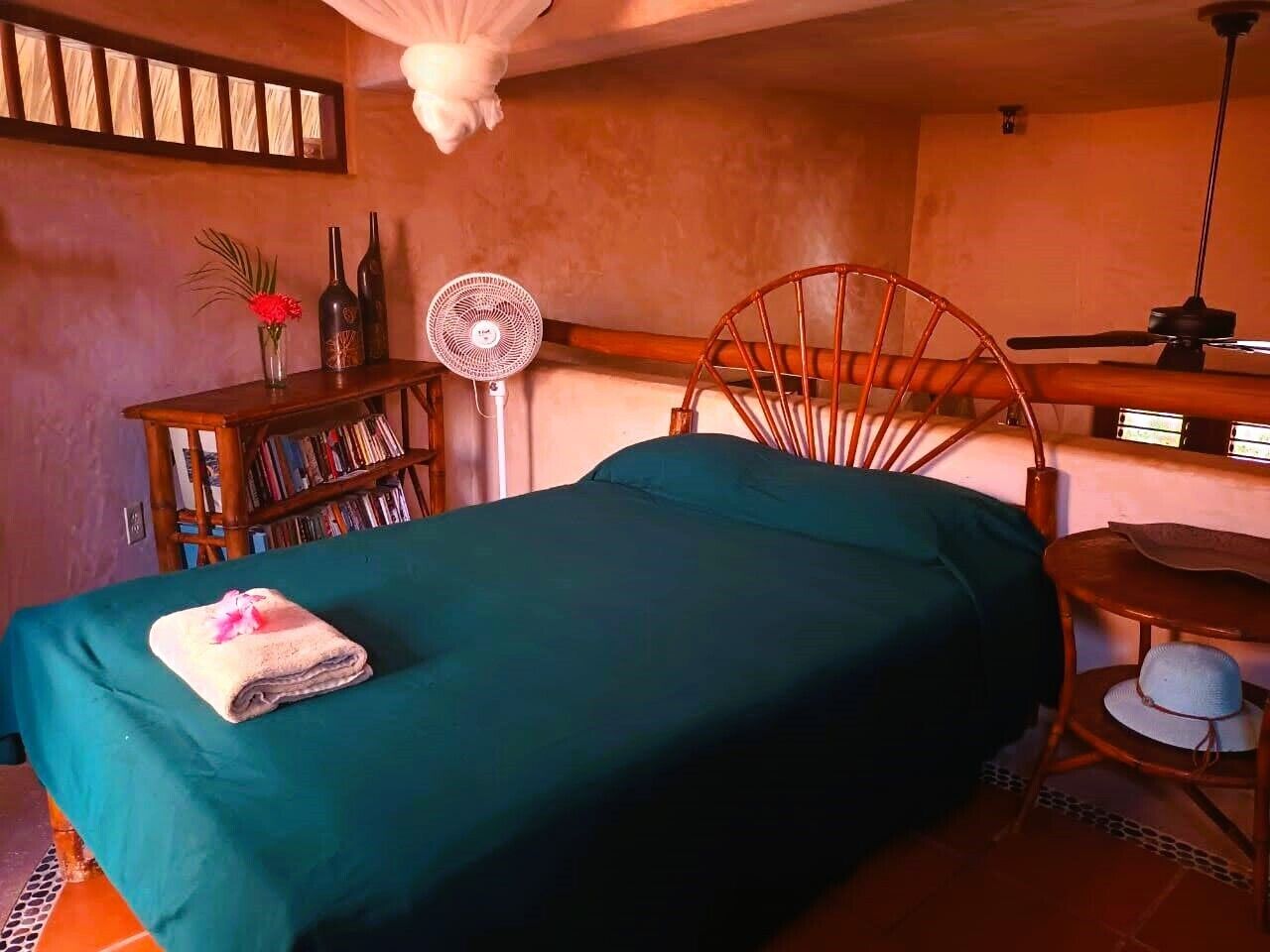 Un dormitorio con cama, una silla de madera, una mesita, una estantería y un ventilador.