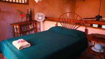 Un dormitorio con cama, una silla de madera, una mesita, una estantería y un ventilador.