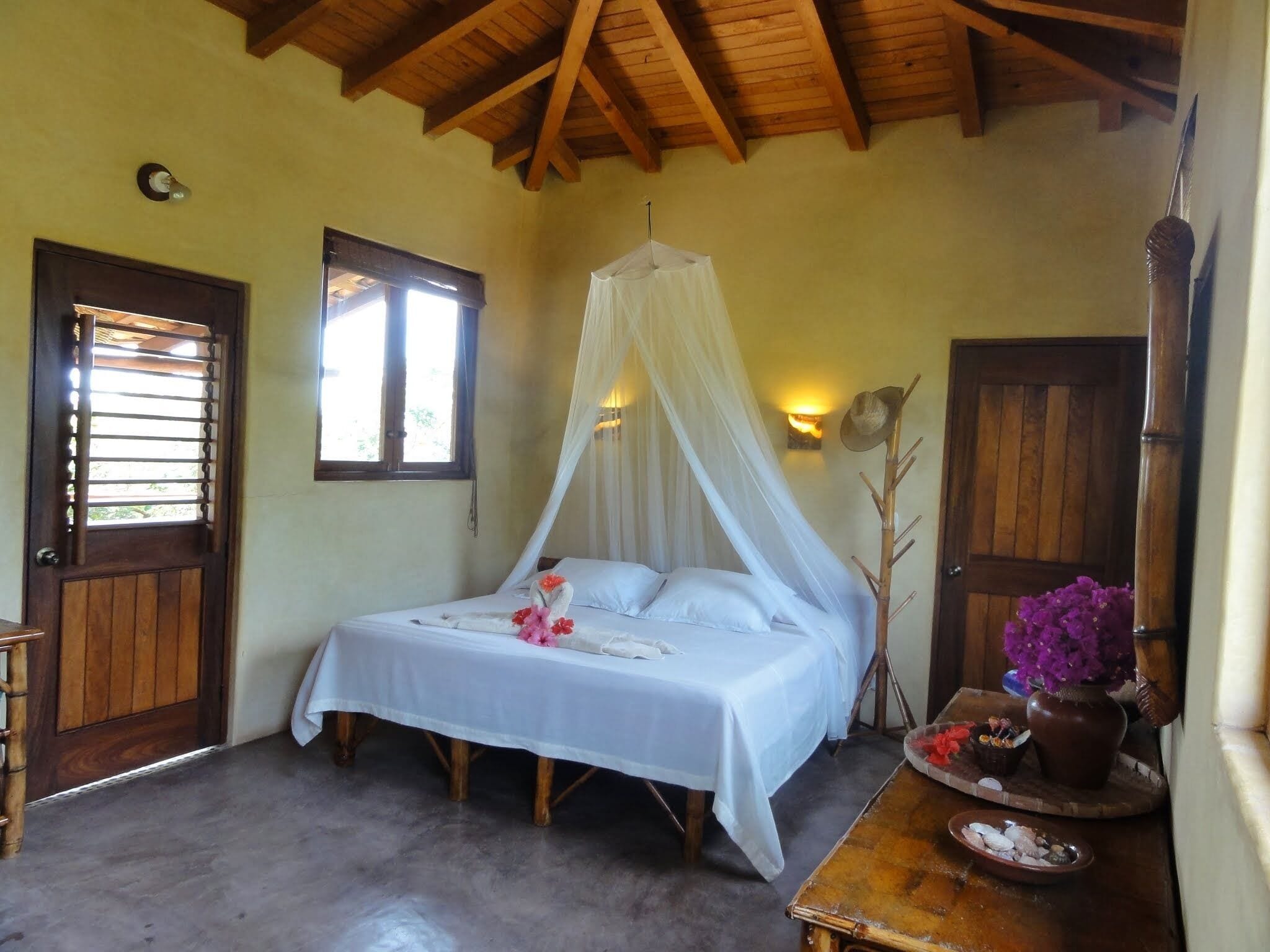 A room with a bed under a canopy, a wooden door, a small table with a vase of flowers, and a wall-mounted clock.
