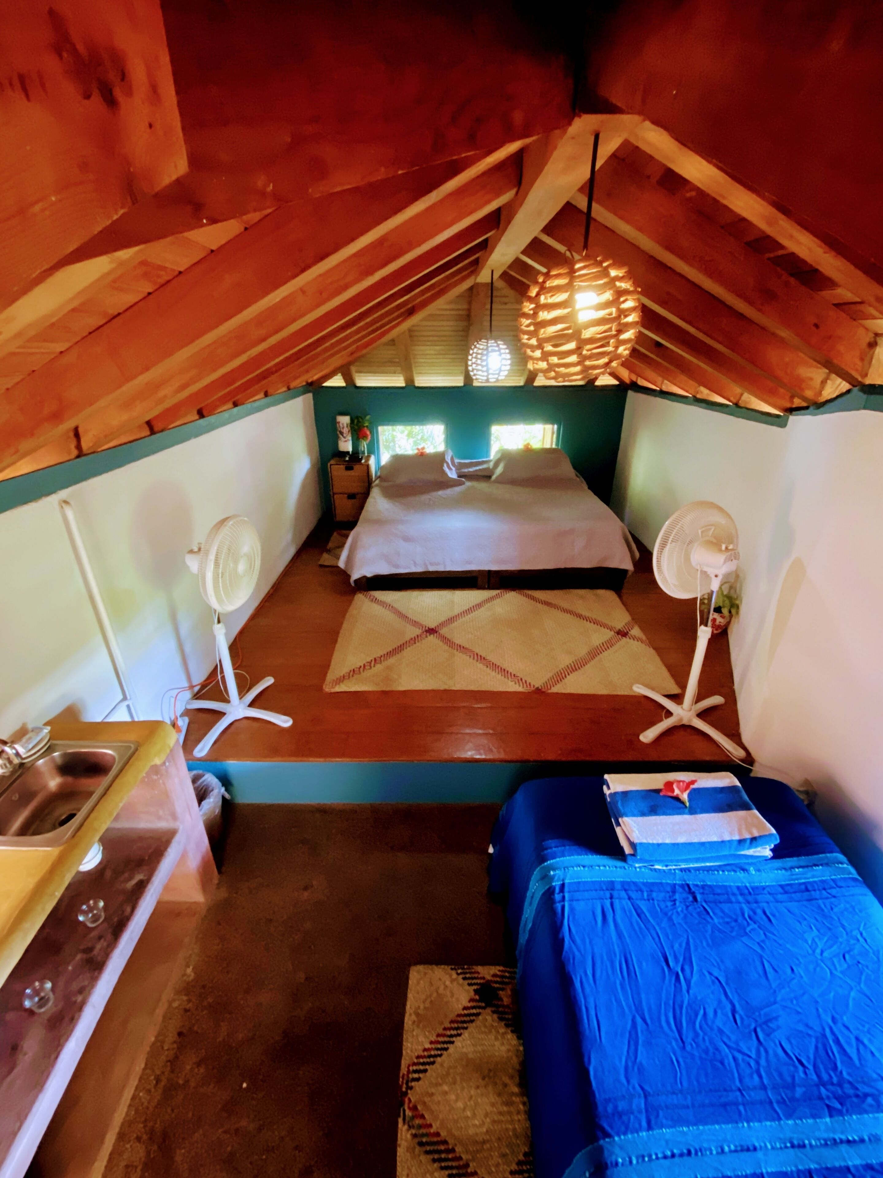 A cozy attic bedroom with a bed, two fans, a small table with a sink, and a window.