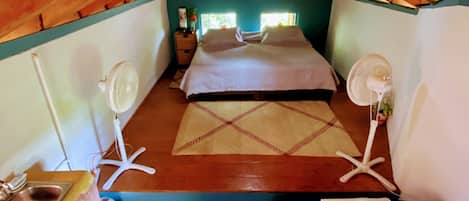 Een gezellige slaapkamer onder de dakpannen met een bed, twee ventilatoren, een klein tafeltje met een wastafel en een raam.