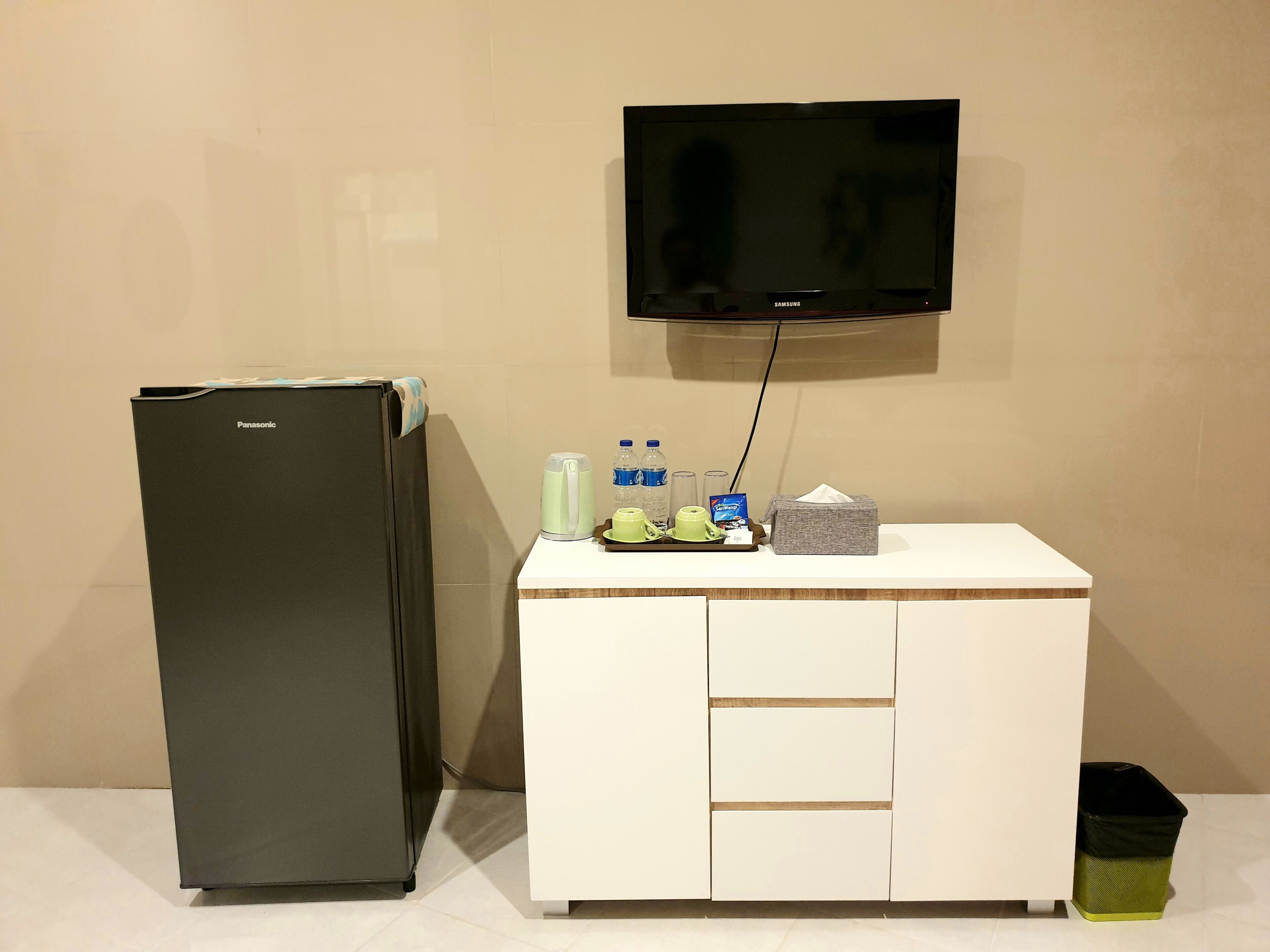 A compact kitchenette with a refrigerator, a small table with a television, and basic amenities like a kettle, water bottles, and a tissue box.
