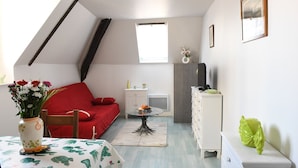 A small, well-lit room with a red sofa, a dining table with a cactus-patterned tablecloth, a small side table, a white cabinet, and a window with a view of trees. - Le Verdier (Dieppe)