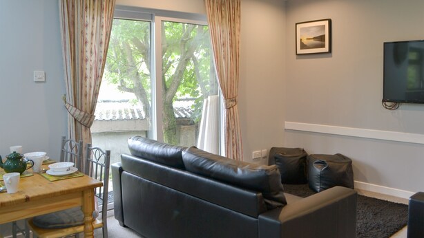 A living room with a sofa, a dining table with chairs, a television mounted on the wall, and a window with curtains. - 3 bedroom accommodation in Oban (Oban)