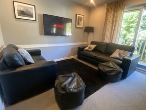 A modern living room with two black sofas, a black ottoman, a flat-screen TV mounted on the wall, and a large window with curtains.