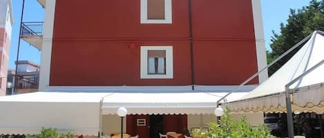 Un edificio de dos pisos con fachada roja, detalles blancos y un área de estar al aire libre cubierta con mesas y sillas.