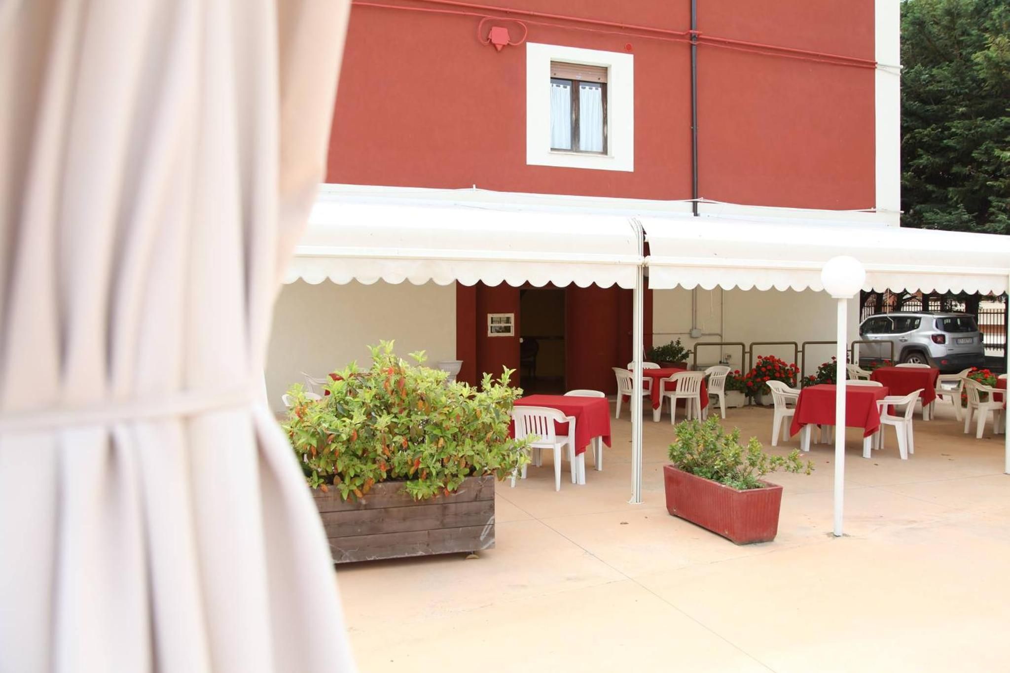 Un'area patio all'aperto con tavoli e sedie, un edificio rosso sullo sfondo e piante in vaso.