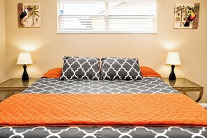 Cama com colcha laranja, duas almofadas estampadas, janela com persianas e duas obras de arte na parede.