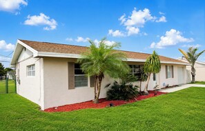 A single-story house with a sloped roof, a front lawn, and a small garden area with red mulch and palm trees. - Centrally located 3Br2Ba Pool Home available for LONGER STAYS, Monthly Discounts (Port St. Lucie)