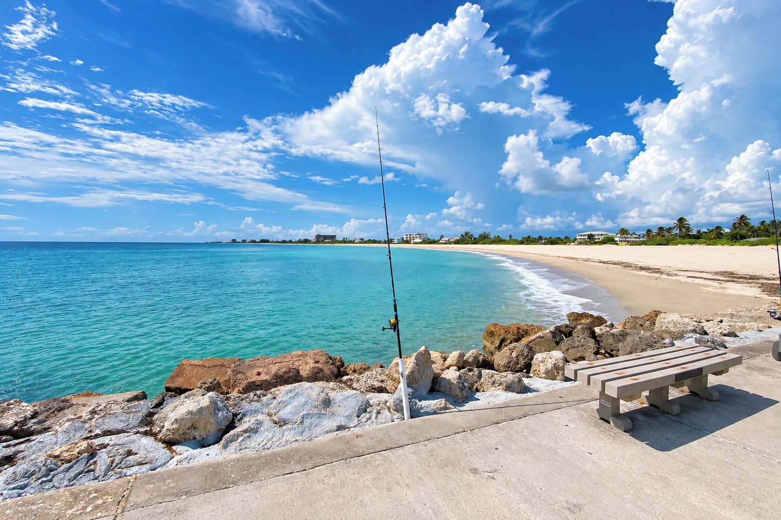 A beach with clear turquoise water, a rocky shoreline, and a concrete walkway with a bench.