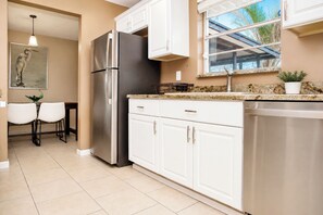 A kitchen with white cabinets, a stainless steel refrigerator, and a dishwasher. There is a window with a view of palm trees.