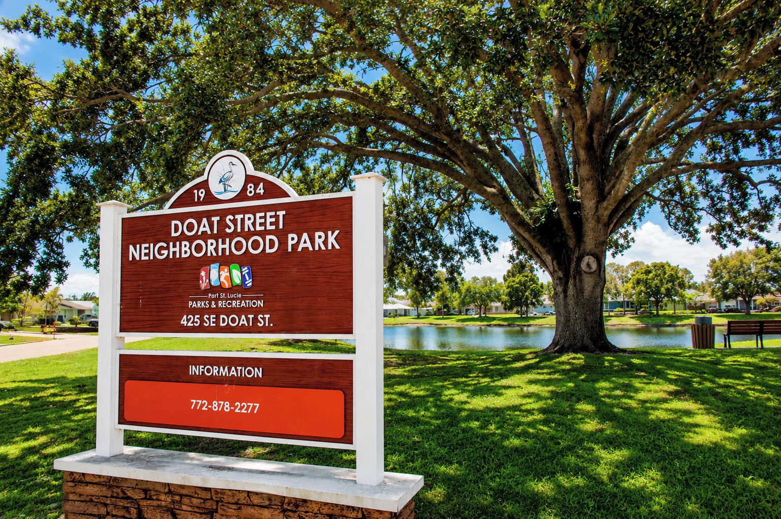 A neighborhood park sign with a large tree and a body of water in the background.