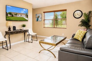 A living room with a TV, a desk, a glass coffee table, a leather sofa, and a window with blinds.