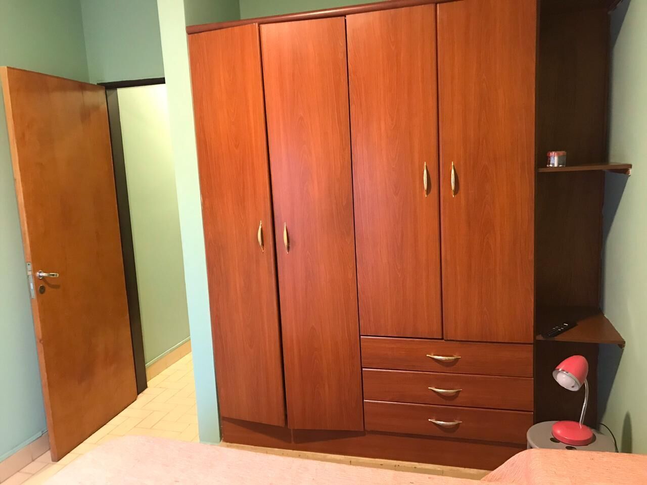A room with a wooden wardrobe, a small desk, and a pink lamp.