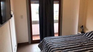 Un dormitorio con cama, televisión montada en la pared, ventana con cortinas y mesita de noche con lámpara.