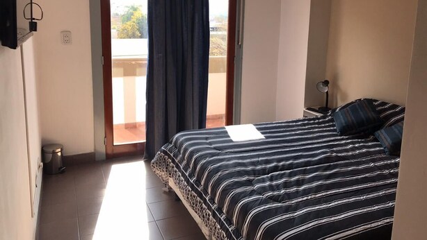 Un dormitorio con cama, mesita de noche, lámpara, ventana con cortinas y vistas al exterior.