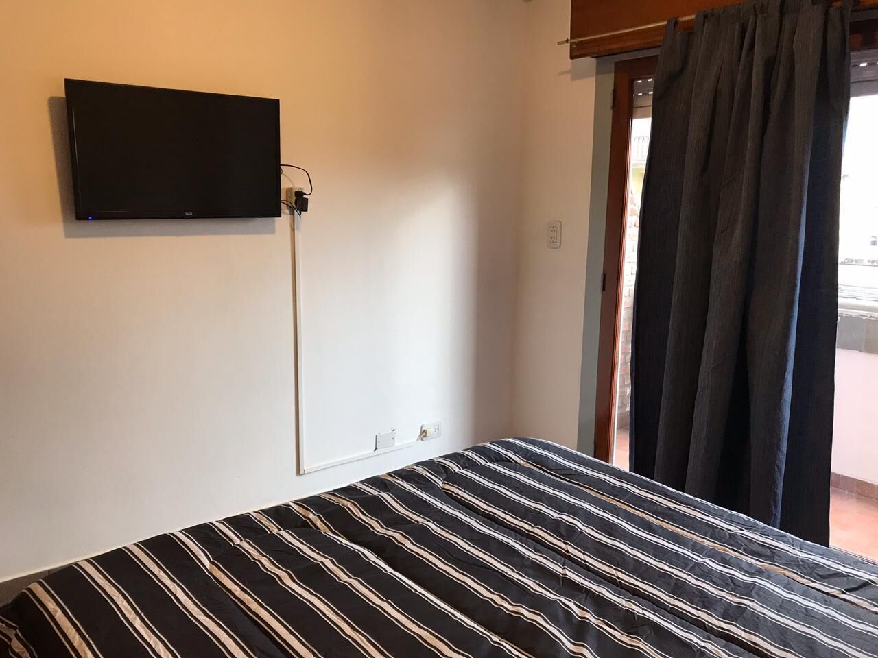 Une chambre avec un lit, un téléviseur fixé au mur et une fenêtre avec des rideaux.