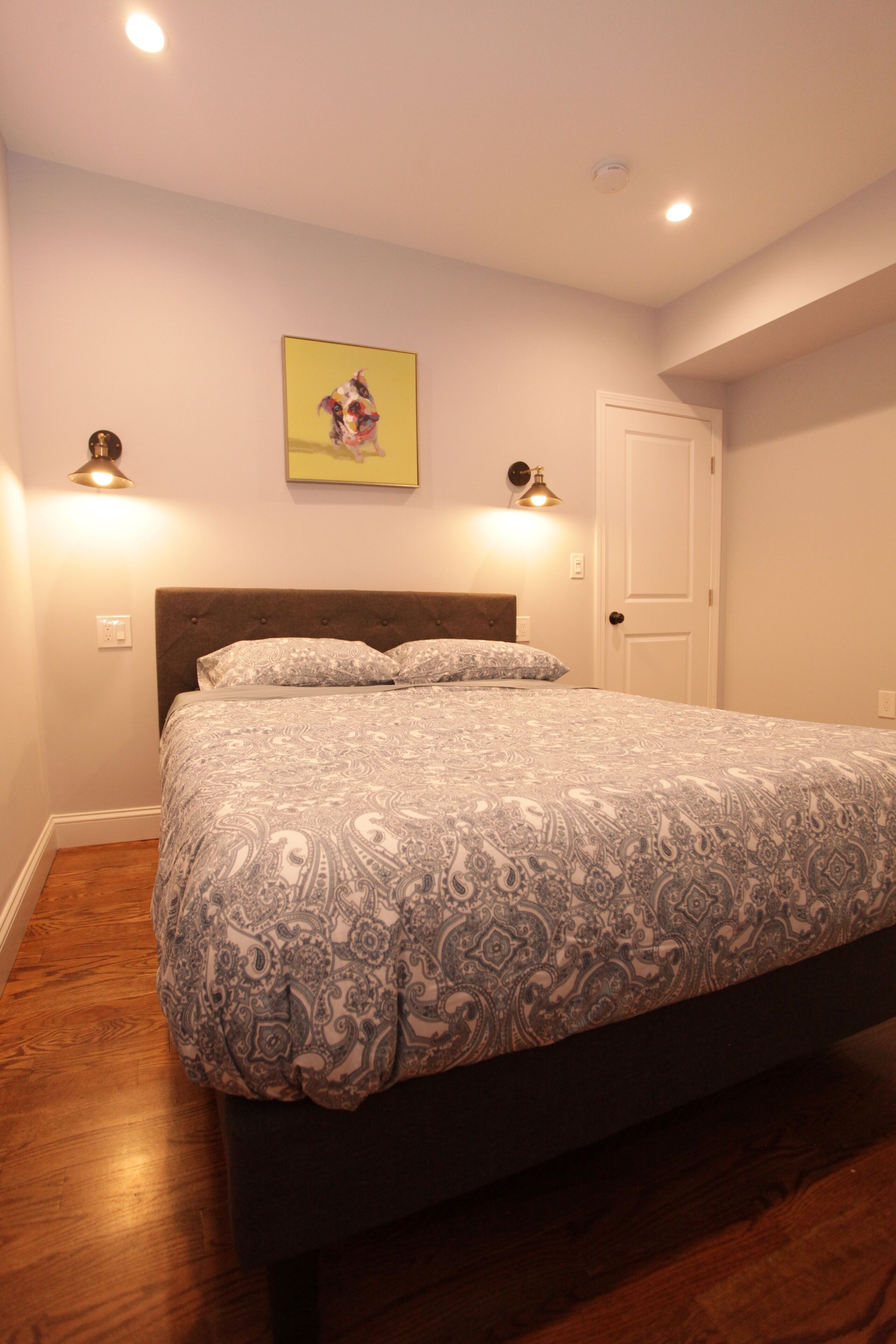 A bedroom with a bed, wooden flooring, a door, and wall-mounted lights.