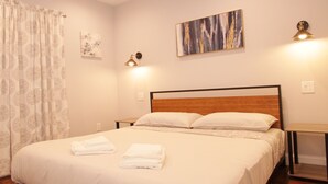 Una cama bien hecha con sábanas blancas, cabecera de madera y mesita de noche con lámpara.