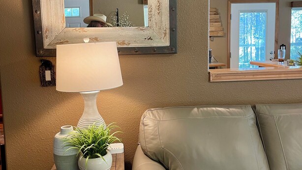 Sala de estar com sofá de couro bege, mesa lateral de madeira com abajur e objetos decorativos, espelho grande com moldura desgastada e vista para o exterior pela janela.