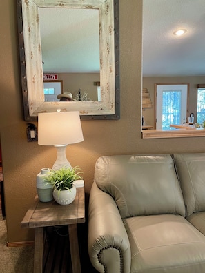 A living room with a beige leather sofa, a wooden side table with a lamp and decorative items, a large mirror with a distressed frame, and a view of the outdoors through a window. - Cabin w/ Hot Tub, 2 Car Garage, close 3 min walk to the Lake! (Island Park)