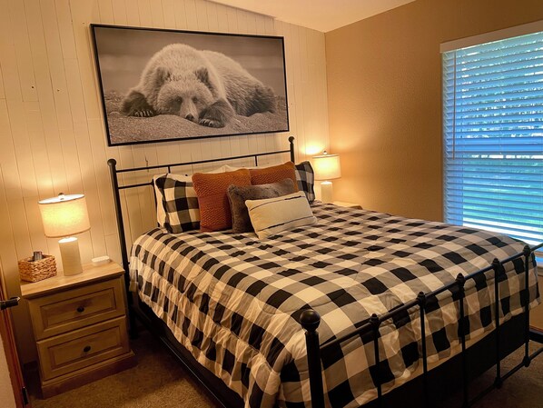 A bedroom with a checkered bedspread, two bedside lamps, a framed picture on the wall, and a window with blinds. - Cabin w/ Hot Tub, 2 Car Garage, close 3 min walk to the Lake! (Island Park)