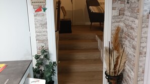 Un couloir avec un plancher en bois, une porte ornée d’une pancarte « BIENVENIDO », une plante en pot et un vase décoratif contenant des plumes d’herbe des pampas.