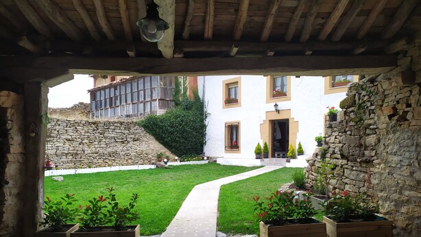 Une cour bordée d’un mur de pierre, agrémentée d’une pergola en bois et d’un chemin menant à un bâtiment à la façade blanche et aux nombreuses fenêtres.