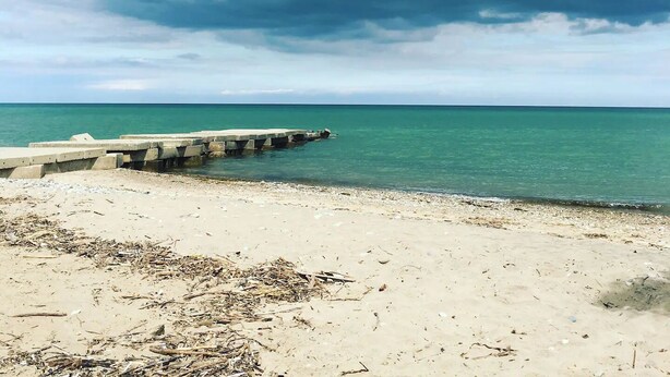 Une plage avec une digue en béton qui s’avance dans la mer, un rivage sablonneux et un ciel nuageux.