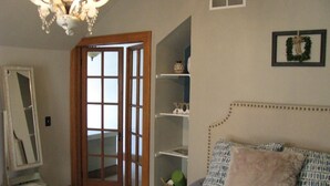 Une chambre à coucher comprenant un lit, une commode, un miroir, un lustre, une porte et une étagère ornée d’objets décoratifs.