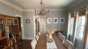 Une salle à manger avec une table en bois, des chaises à revêtement blanc, un lustre et une bibliothèque en bois.
