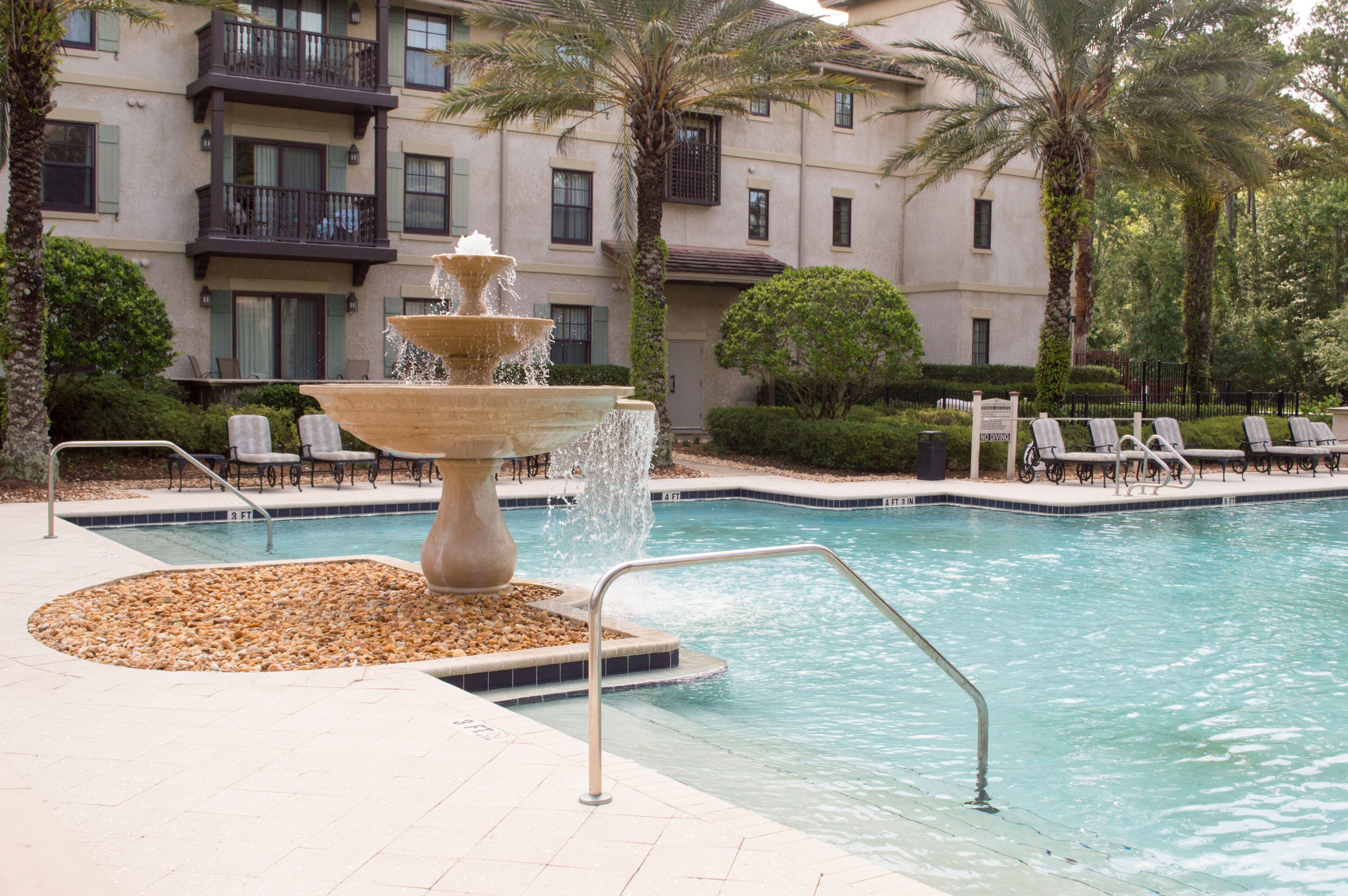 A swimming pool with a tiered water fountain, lounge chairs, and a building with balconies in the background.