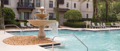 A swimming pool with a tiered water fountain, lounge chairs, and a building with balconies in the background.