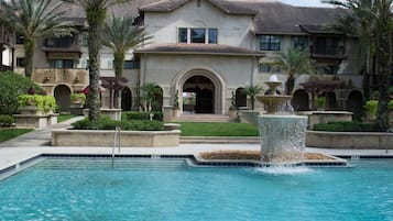 A swimming pool with a fountain, surrounded by palm trees and a large building with multiple balconies.