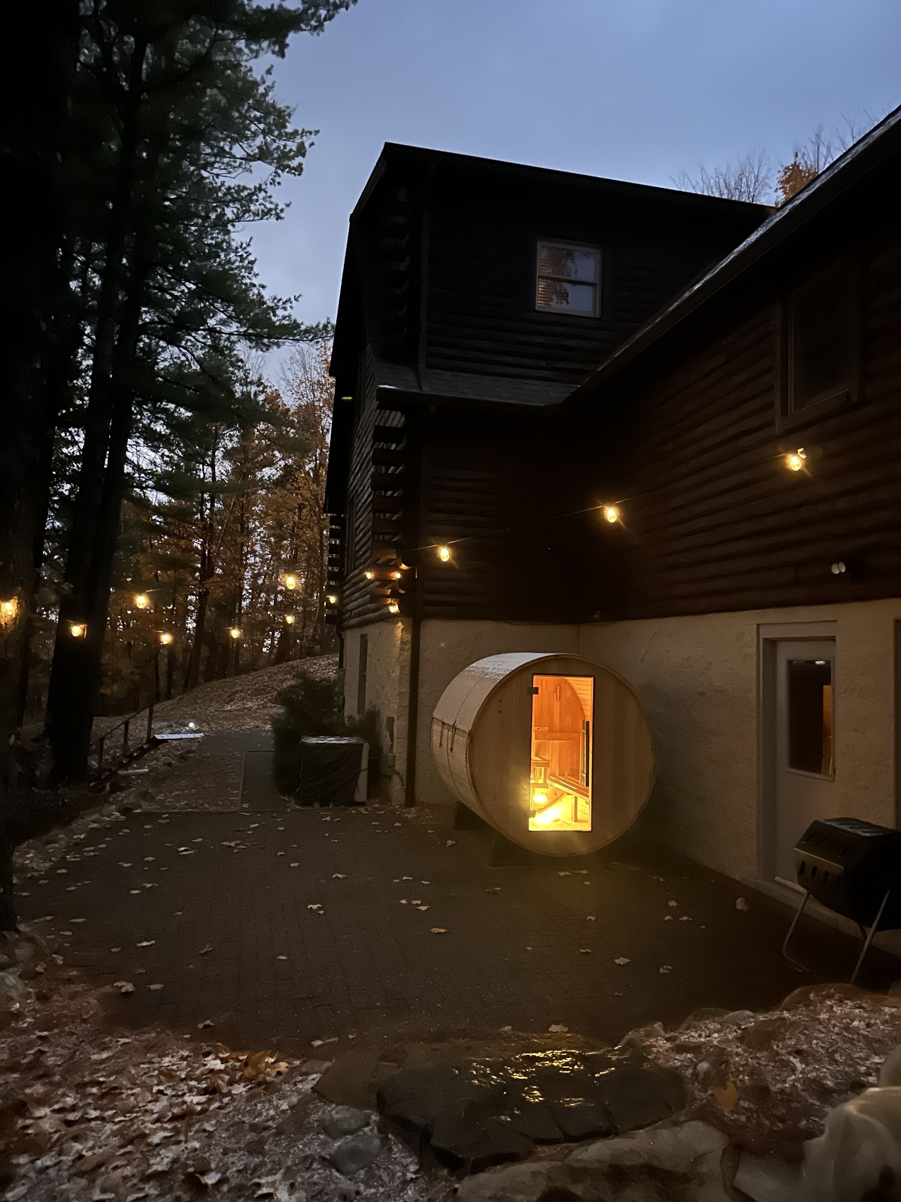 Una cabaña de troncos con una estructura cilíndrica grande adosada, una terraza de madera y luces colgantes.