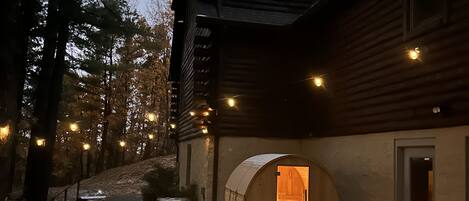 Una cabaña de troncos con una estructura cilíndrica grande adosada, una terraza de madera y luces colgantes.