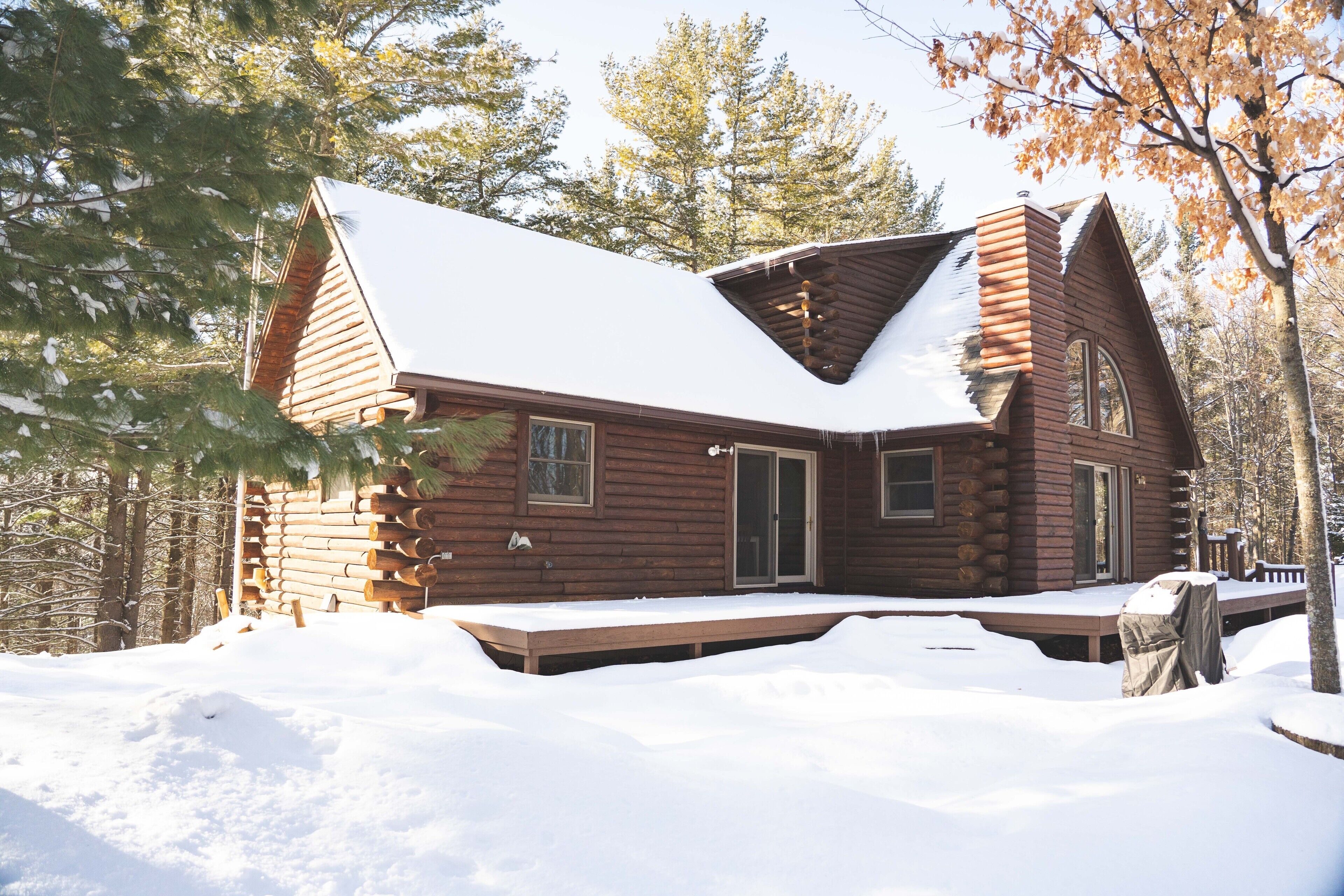 A log cabin with a snow-covered roof, a deck, and a forested backdrop.