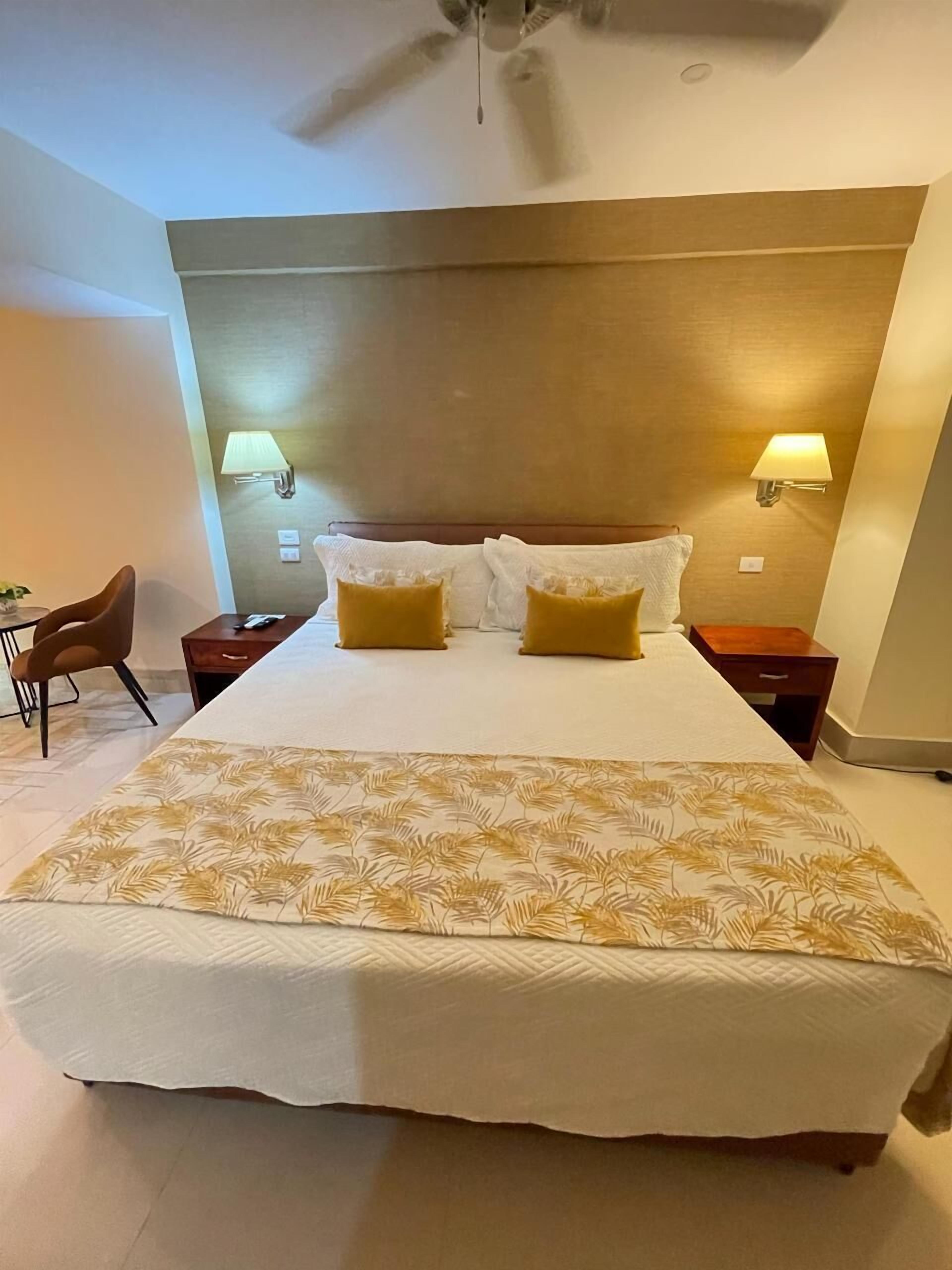 A hotel room with a large bed, two bedside lamps, a ceiling fan, and a small table with a chair.