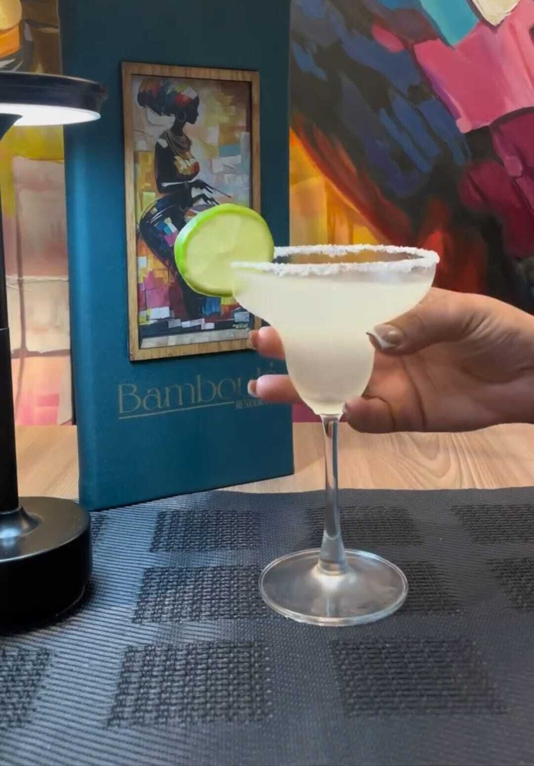 En hand håller i en margarita med en limeskiva, bredvid står ett inramat konstverk och en bok med titeln ”Bambou” på ett bord.