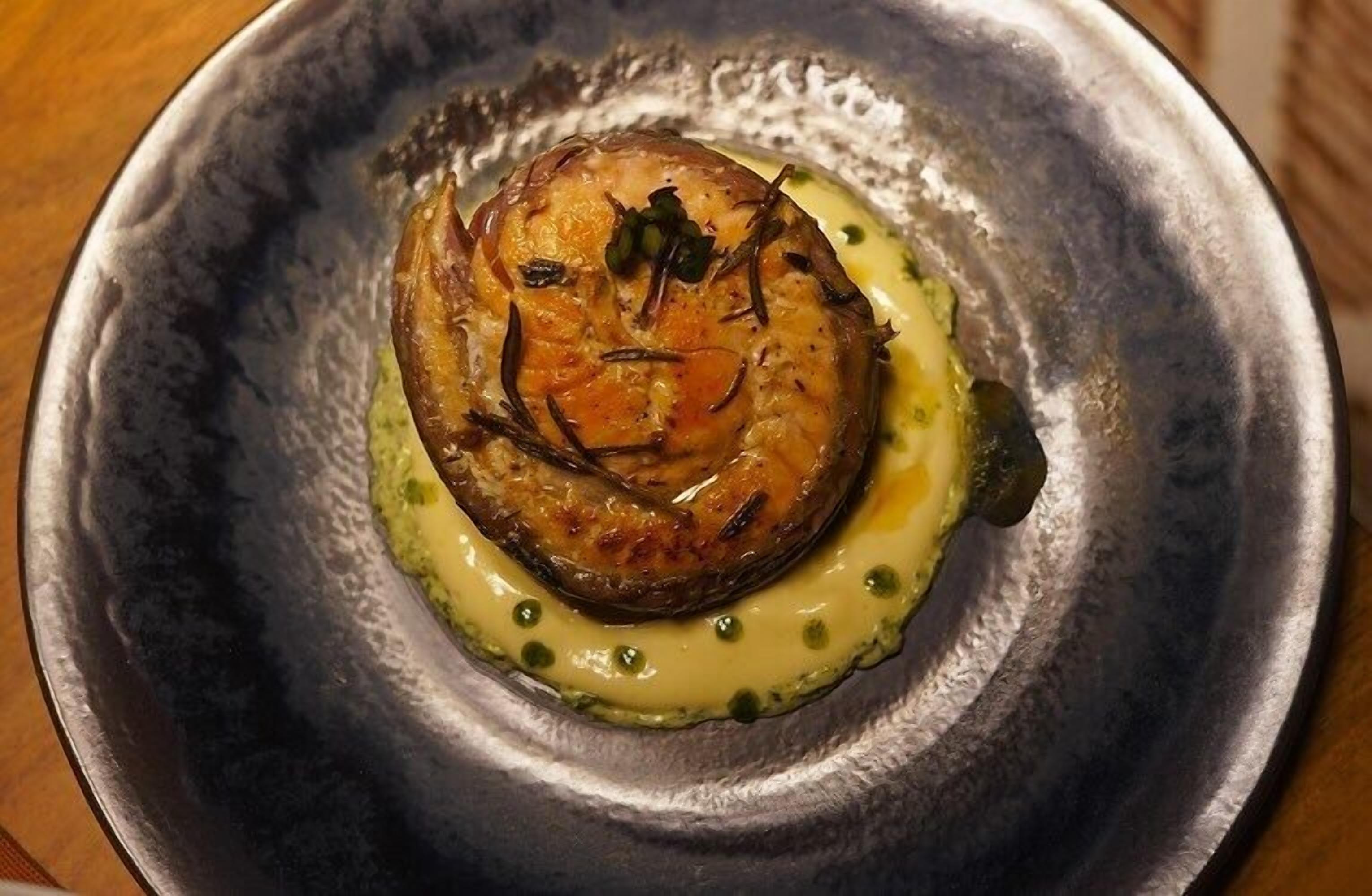 A dish with a round, golden-brown center, garnished with herbs, on a dark plate with a green sauce around the edge.
