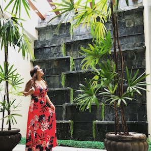 Una mujer con un vestido floral parada en un patio con plantas en macetas y un muro de piedra.