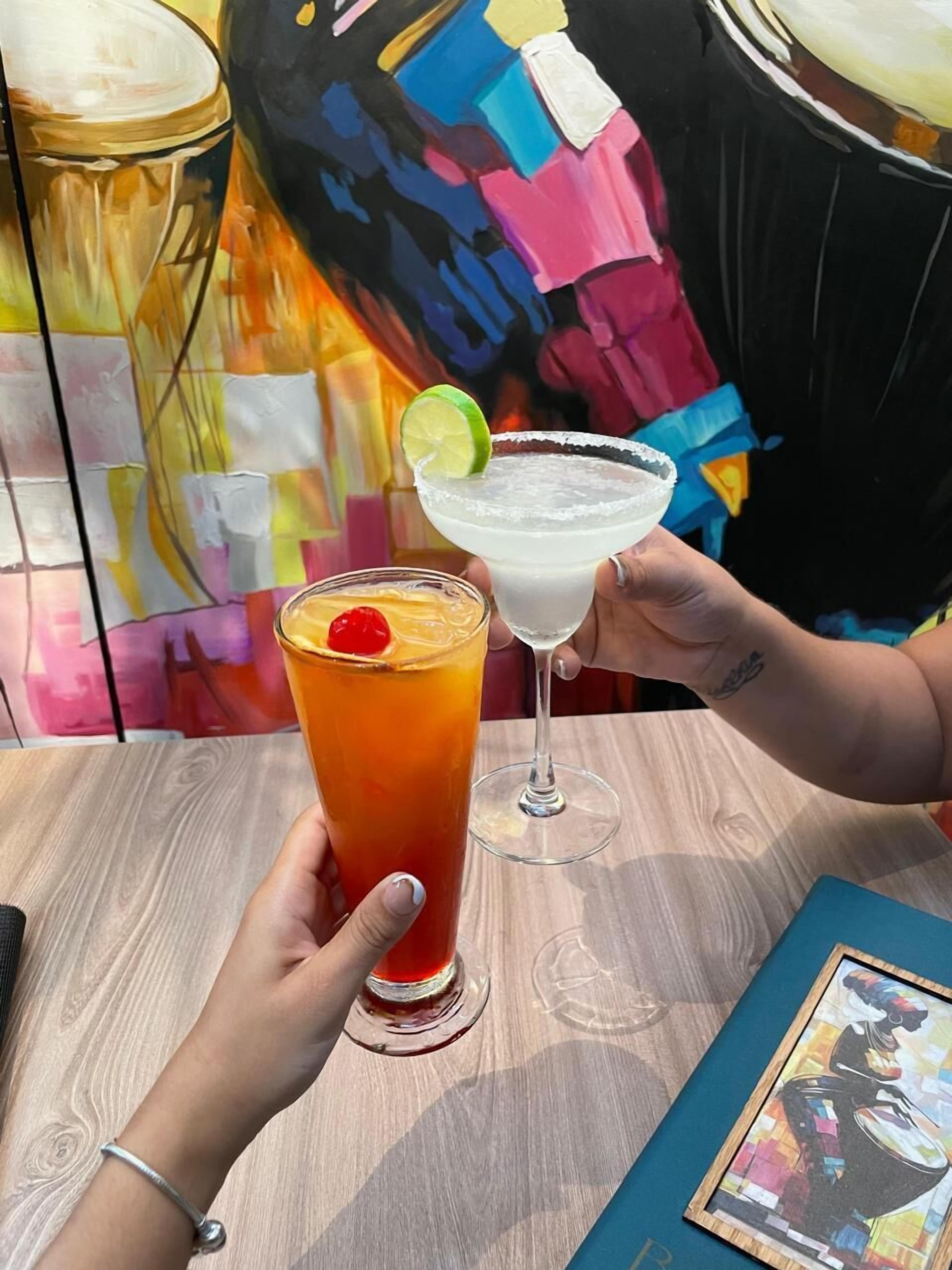 Two drinks, one in a tall glass and one in a margarita glass, held by two people at a wooden table with a colorful painting in the background.