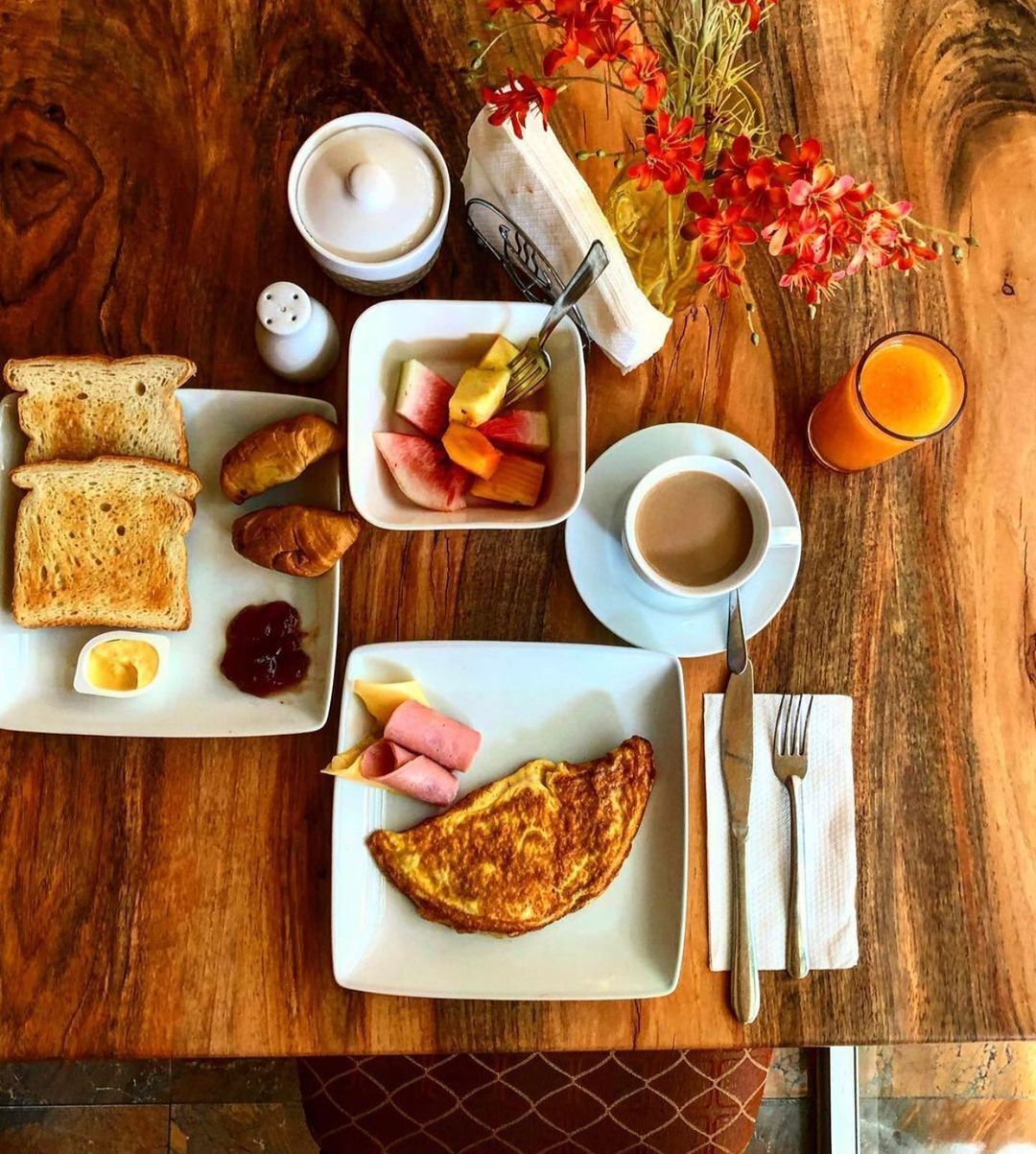 Desayuno con tostadas, croissants, jamón, huevos, fruta, café y jugo sobre una mesa de madera.