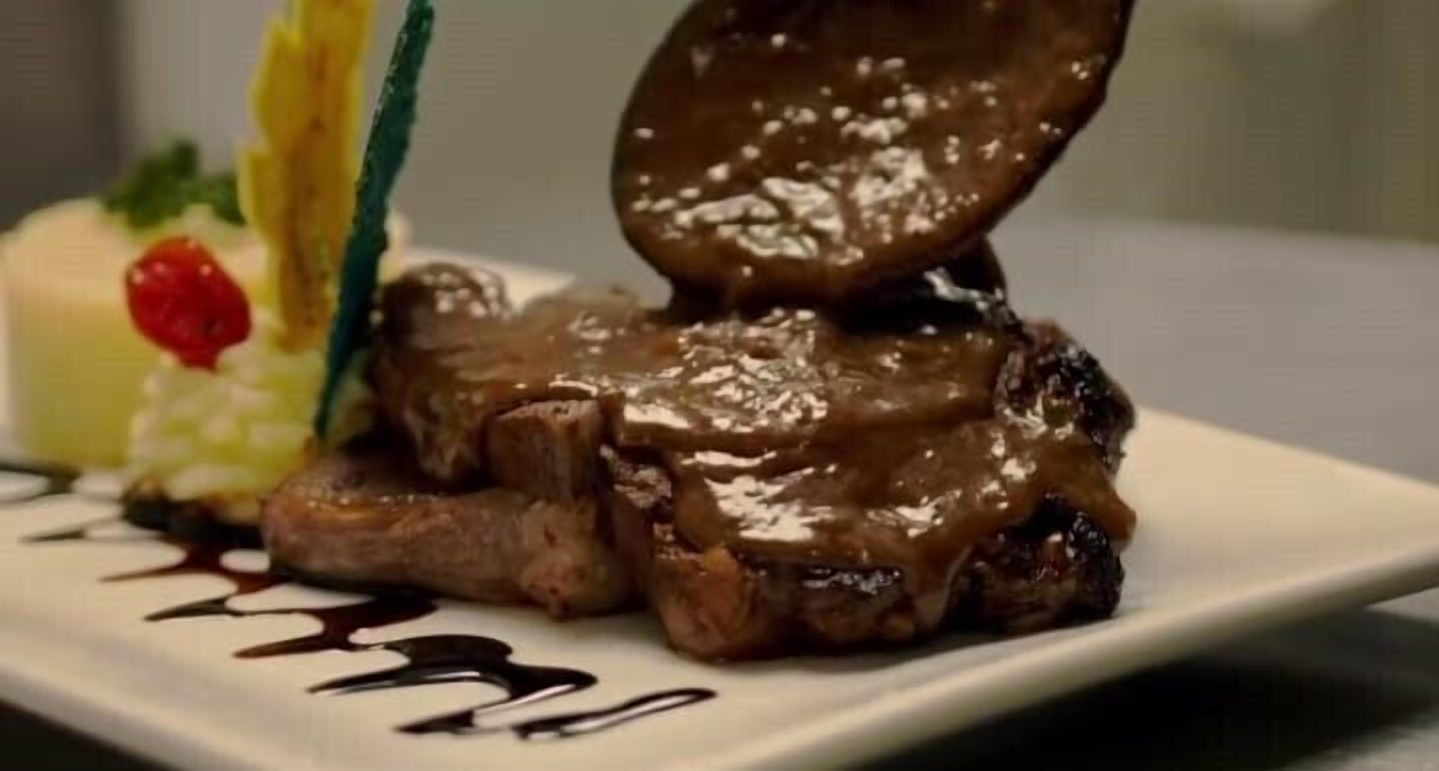 A plate with a steak topped with a brown sauce, accompanied by a small dessert and a decorative element.