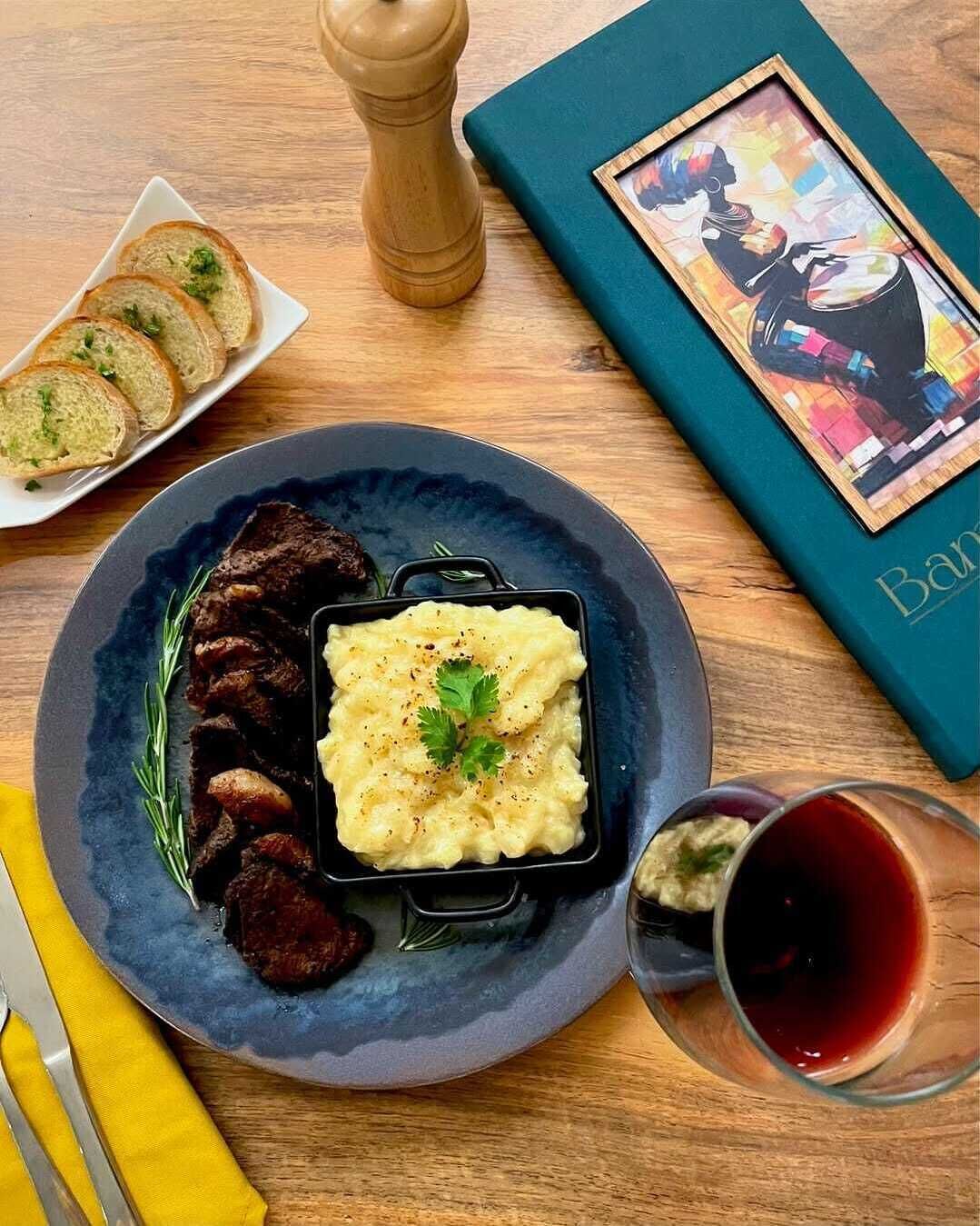 A plate with mashed potatoes and meat, a glass of red wine, and a book on a wooden table.