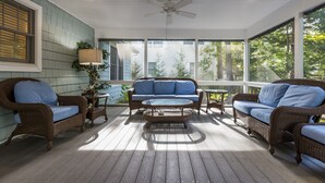 Eine überdachte Veranda mit Korbmöbeln, einem Deckenventilator und großen Fenstern mit Blick auf Bäume.