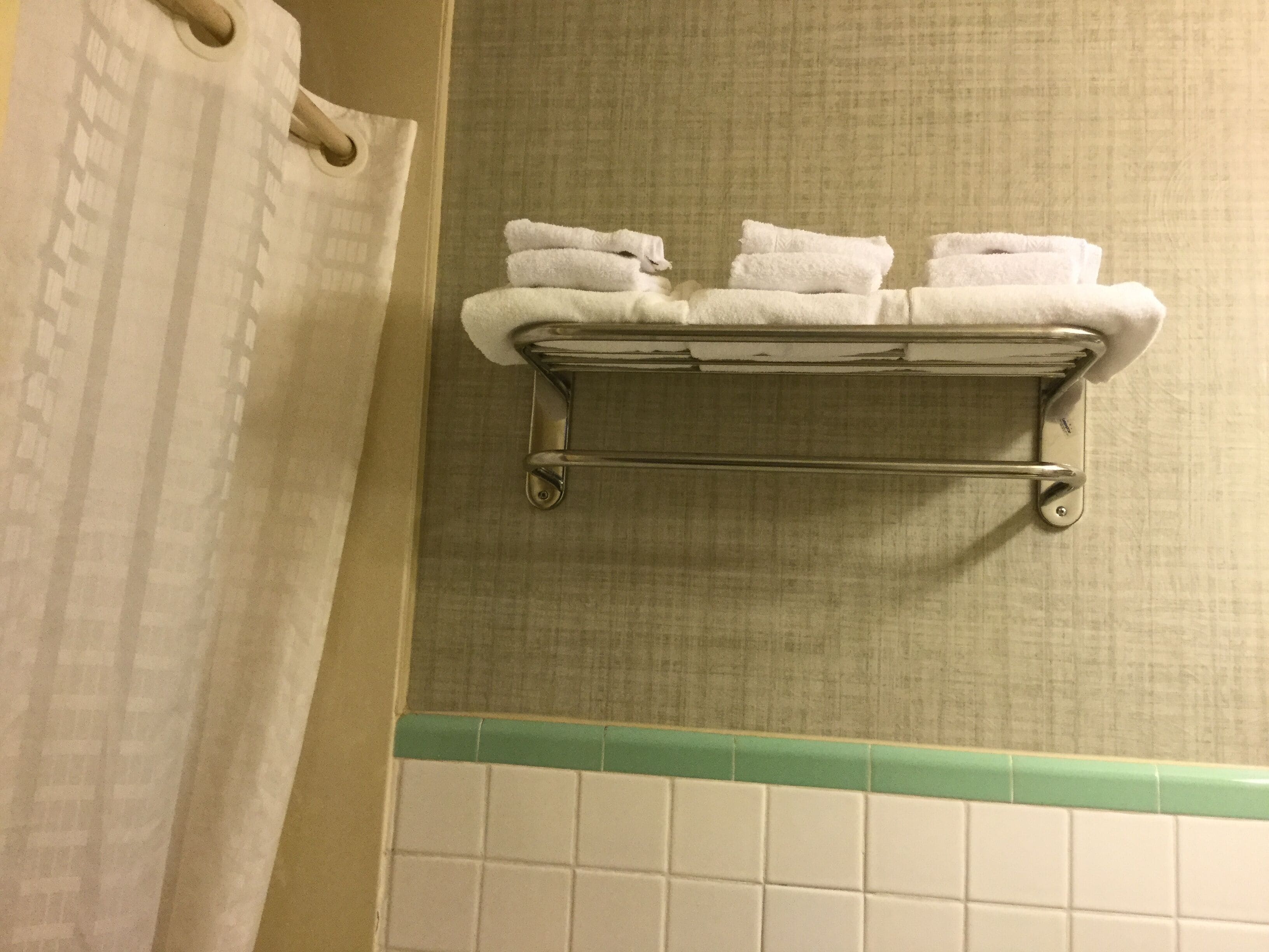 A bathroom with a towel rack holding three folded towels, a shower curtain, and a green-tiled wall.